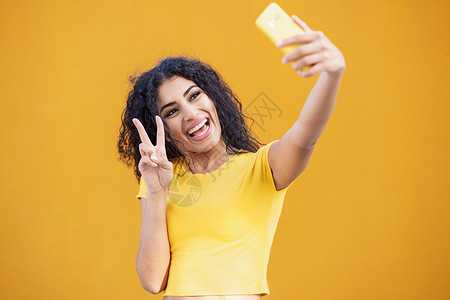 有趣的阿拉伯女人用智能手机拍自拍照片卷发女性年轻的阿拉伯女人用智能手机自拍黄色的高清图片素材