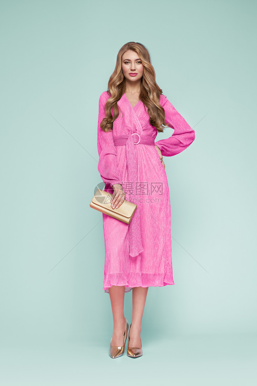 穿着优雅粉红色连衣裙的金发年轻女人模型摆蓝色背景上的全长珠宝发型带手提包的女孩时尚照片图片