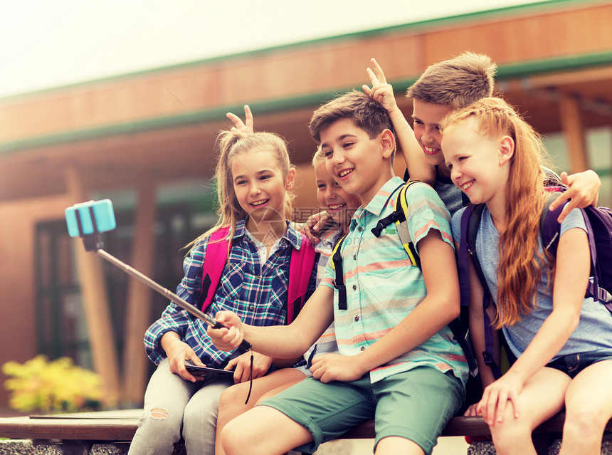 小学教育,技术,友谊,童年人的群小学生着背包坐长凳上,用智能手机户外自拍杆上拍照快乐的小学生自拍图片