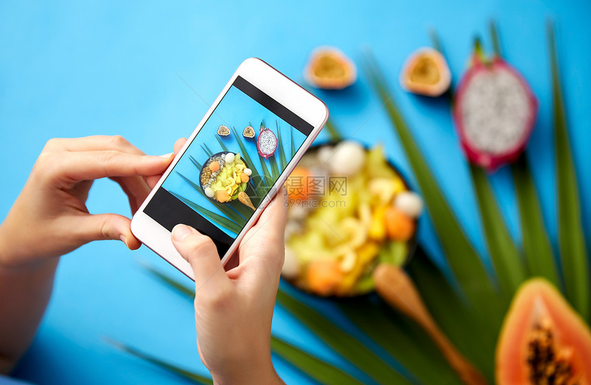 食物,技术健康的饮食手与智能手机拍照同的异国水果混合碗与木制勺子绿色风扇棕榈叶蓝色背景双手智能手机上拍摄异图片