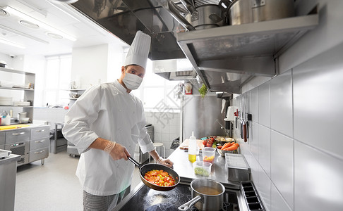健康安全大流行男厨师戴着面部防护医学面具,餐厅厨房的煎锅中防止病疾病烹饪食物餐厅厨房面具烹饪食物的厨师背景图片