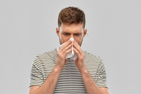 鼻炎,感冒过敏的年轻人用纸巾吹他的鼻子灰色的背景年轻人用纸巾吹鼻子图片