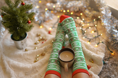 圣诞树附近穿着圣诞袜的脚女人坐毯子上,喝热饮料,放松,穿着羊毛袜子热身寒假圣诞节的背景图片