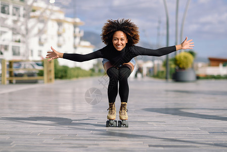 年轻合身的黑人妇女溜冰鞋骑户外的城市街道上张开双臂阳光明媚的日子里,微笑的女孩留着AFRO的发型背景图片