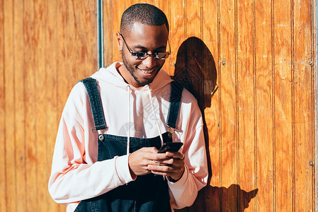 年轻的黑人男子穿着休闲服装太阳镜,木制背景下用智能手机千禧年的非洲人带着围嘴裤户外微笑背景图片