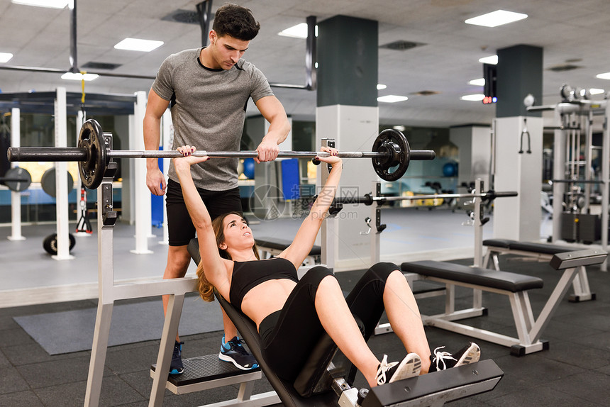 私人教练帮助个年轻的女人健身房锻炼时举重图片