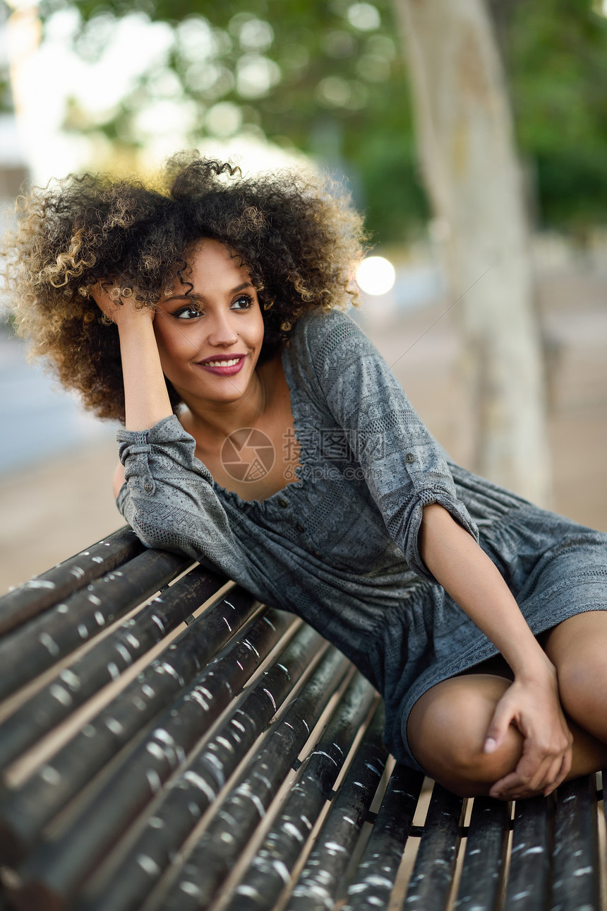 有趣的黑人女人,留着AFRO发型,坐城市背景的长凳上穿着休闲衣服的混合女孩年轻的黑人妇女,城市背景图片