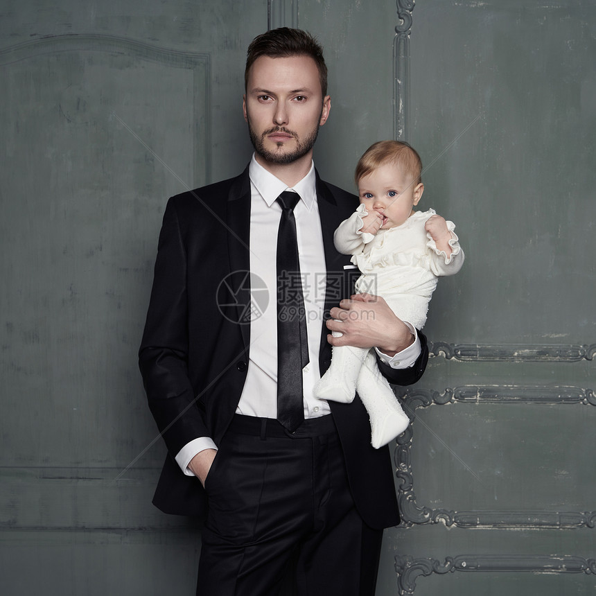 年轻的父亲怀里抱着漂亮的小婴儿有漂亮女儿的男人的时尚肖像时尚的爸爸晚上黑色西装小女孩穿着白色滑块工作室肖像图片
