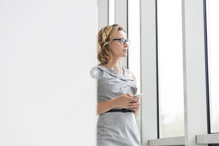 有思想的女商人着智能手机,站新办公室的窗边图片