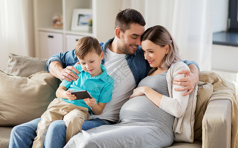 家庭,怀孕技术快乐的怀孕母亲,父亲小儿子坐家里的沙发上智能手机家里智能手机的幸福家庭人高清图片素材