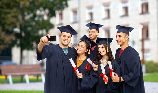 毕业生自拍教育,毕业人的群快乐的研究生砂浆板学士学位礼服与文凭采取自拍智能手机校园建筑背景毕业证书的毕业生用手机自拍背景