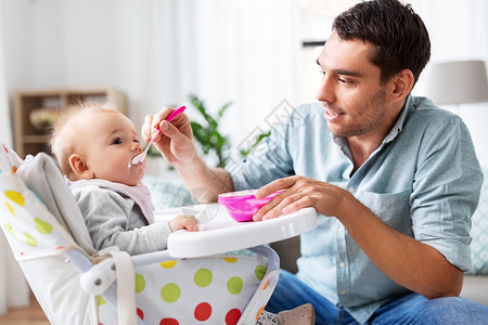 家庭,食物,饮食人的快乐的父亲喂养小婴儿女儿坐高椅子上,用勺子家里父亲家高椅子上喂快乐的婴儿晚餐高清图片素材