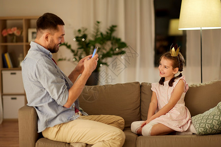 家庭技术人的快乐的父亲晚上家用智能手机拍摄女儿父亲家用手机给女儿拍照图片