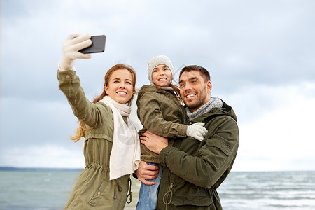 养育子女照片家庭休闲人的快乐的母亲父亲小女儿秋天的海滩上用智能手机自拍家人秋天的海滩上用智能手机自拍背景