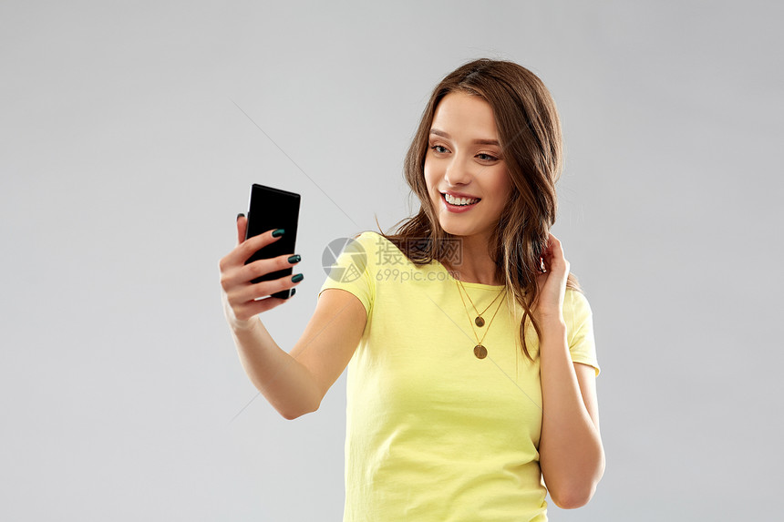 技术人的微笑的轻妇女十几岁的女孩穿着空白的黄色T恤,用智能手机灰色背景下自拍微笑的十几岁女孩用智能手机自拍图片