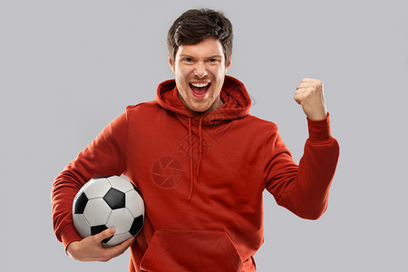 球帽运动,休闲游戏获胜快乐的人足球迷穿着红色帽衫,带着足球庆祝胜利,并灰色背景上拳泵手势快乐的人足球迷,足球获胜背景