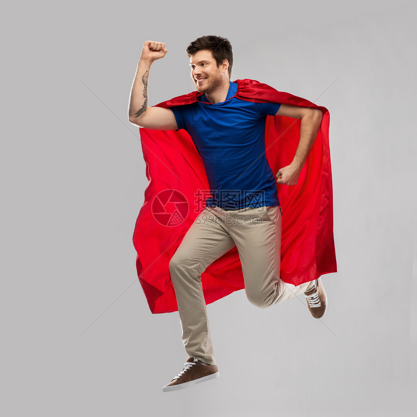 超级力量人的快乐的轻人穿着红色超级英雄斗篷灰色背景下空中跳跃穿着红色超级英雄斗篷的男人空中跳跃图片