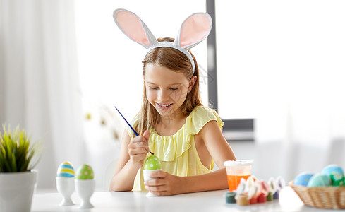 叼着铅笔的兔子复活节,假日人们的快乐的女孩戴着兔子耳朵,头带着色鸡蛋与颜色刷子家里快乐的女孩家里给复活节彩蛋着色背景