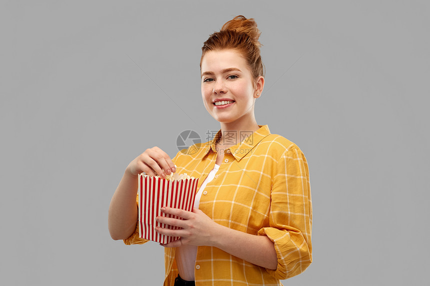 快餐人们的微笑的红色头发十几岁的女孩穿着格子衬衫,吃爆米花条纹桶灰色背景微笑的红头发少女吃爆米花图片