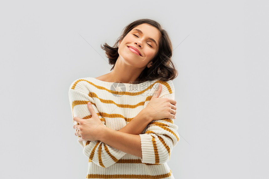 人们的快乐的轻女人穿着条纹套衫,闭着眼睛拥抱自己灰色的背景穿着条纹套衫的快乐女人拥抱自己图片