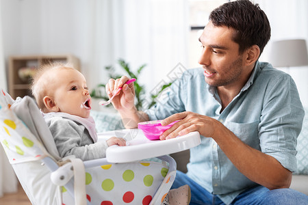 家庭,食物,饮食人的快乐的父亲喂养小婴儿女儿坐高椅子上,用勺子家里父亲家高椅子上喂快乐的婴儿父母身份高清图片素材