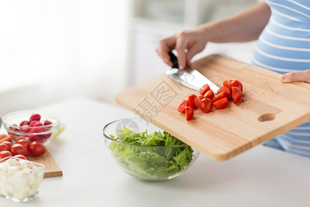 怀孕,烹饪食物健康饮食密切孕妇与菜刀添加切碎樱桃番茄木制切割板沙拉碗家里孕妇家烹饪食物背景图片