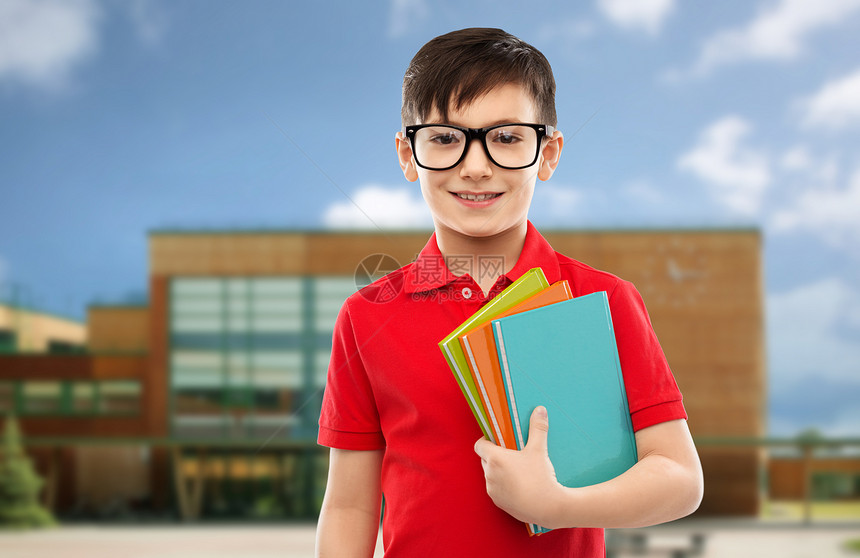 教育人的微笑的小学生男孩穿着红色马球T恤,戴着眼镜,带着学校背景的书带着书戴眼镜的微笑的小学生图片