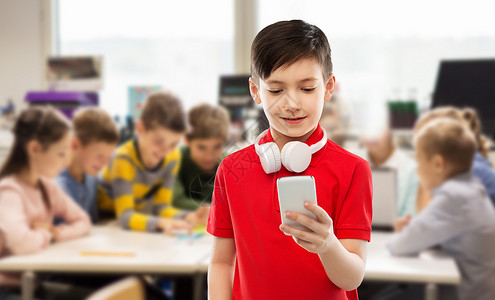 教育技术人的微笑的小男孩穿着红色马球T恤,脖子上戴着耳机,学校的课堂背景上用智能手机带耳机智能手机的红色T恤课程高清图片素材