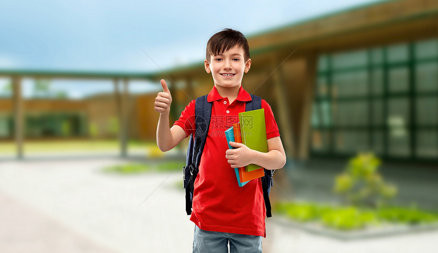 教育人的微笑的小学生男孩穿着红色马球T恤,戴着眼镜,带着书书包,学校背景上竖大拇指学生男孩着书书包竖大拇图片