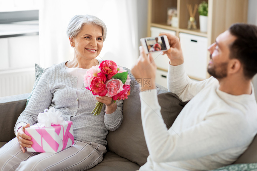 家庭母亲生日微笑的成儿子家里用智能手机拍摄长母亲的鲜花礼品盒成儿子家里给迈的母亲拍照图片