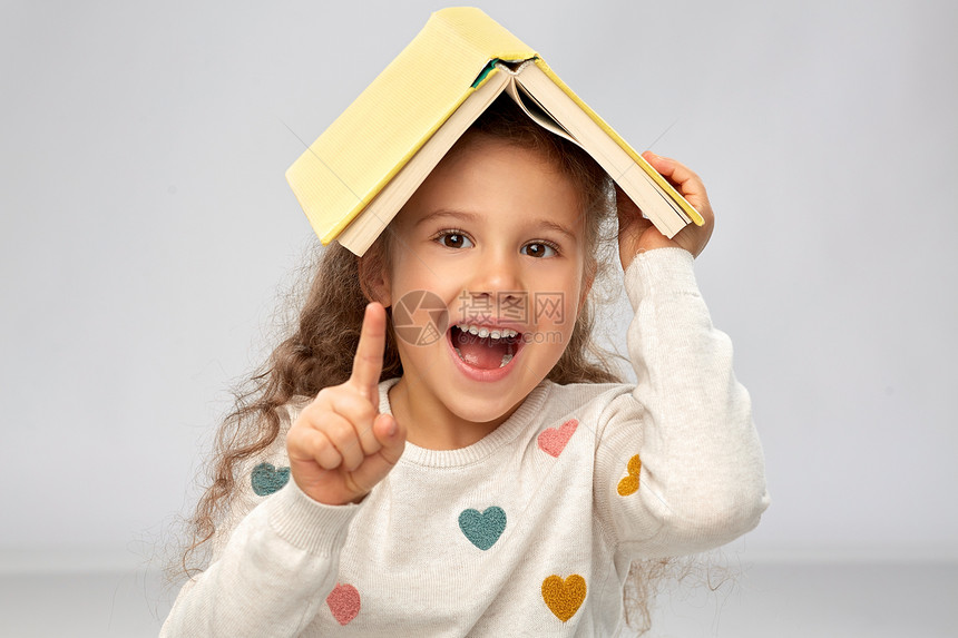 阅读,教育童的微笑的小女孩的肖像,头上书,屋顶上灰色的背景带着书的微笑女孩的肖像图片