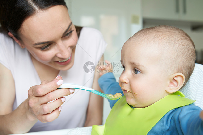 微笑的8个月大的男孩家里的高椅子上被母亲用勺子喂固体食物图片