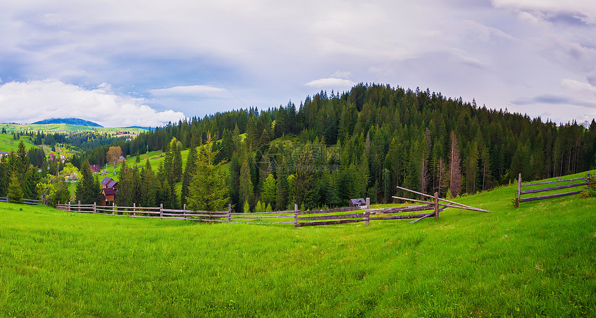 风景如画的春卡皮亚人的场景与木制的分裂铁路围栏跨越个绿色郁郁葱葱的牧场,旧小屋山谷周围的松树林图片
