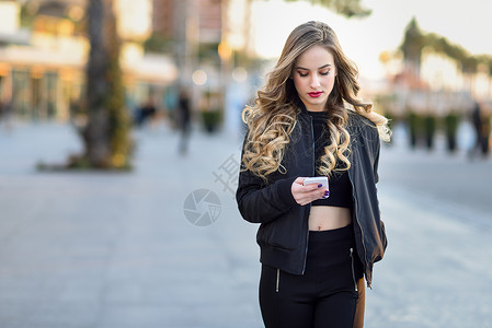 金发女人城市背景下用她的智能手机发短信漂亮的轻女孩穿着黑色夹克街上散步漂亮的俄罗斯女,留着长长的波浪发型背景图片