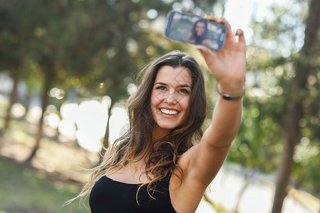 公园里位漂亮的轻女人用智能手机自拍的肖像图片