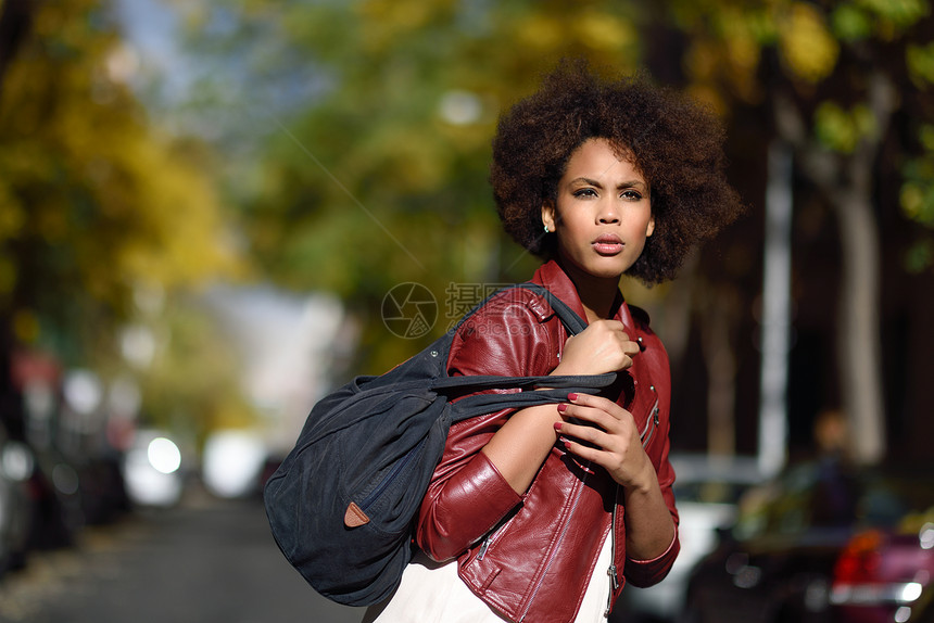 轻的黑人女,留着AFRO发型,站条城市街道上,背着个包混合女人穿红色皮夹克白色连衣裙与城市背景图片