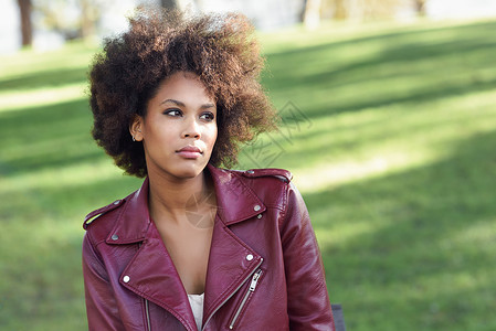 轻的黑人女,留着AFRO发型,坐城市公园的长凳上混合女人穿红色皮夹克白色连衣裙与城市背景背景