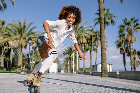 微笑的黑人妇女溜冰鞋骑户外海滩长廊与棕榈树阳光明媚的日子里,微笑的女孩留着AFRO的发型背景图片