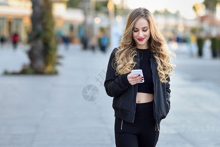金发女人城市背景下用智能手机发短信金发女人城市背景下用她的智能手机发短信美丽的轻微笑的女孩穿着黑色夹克街上散步背景图片