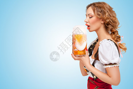 轻口渴感的瑞士女人穿着红色跳线短裤吊带的传统Dirndl的形式,喝啤酒与泡沫流出杯子蓝色背景背景图片