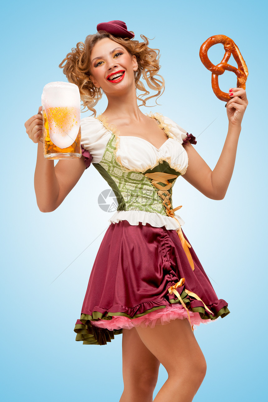 轻感的啤酒节女服务员穿着传统的巴伐利亚连衣裙,着个椒盐卷饼啤酒杯,蓝色的背景下开心地笑着图片