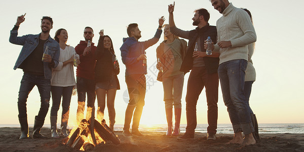 快乐无忧无虑的轻朋友海滩上玩得开心,太阳开始落山的时候篝火旁喝啤酒背景图片