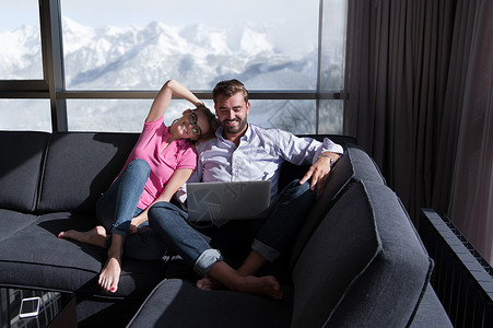 轻的夫妇家里放松,用笔记本电脑客厅里,靠近窗户的沙发上看书图片