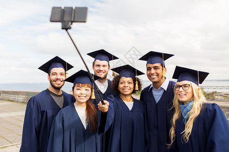 教育毕业技术人的群快乐的国际学生穿着迫击炮板学士服,用智能手机自拍杆户外拍照群快乐的学生毕业生自拍朋友董事会高清图片素材