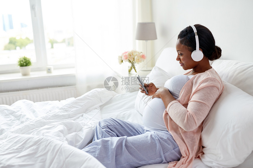 怀孕,技术人的快乐怀孕的非裔美国妇女戴着耳机智能手机家里睡觉带智能手机的耳机里的孕妇带智能手机的耳机里的孕妇图片