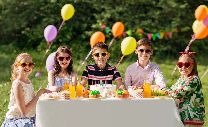 假期,童庆祝快乐的孩子与蜡烛生日蛋糕上坐桌子上的夏季花园聚会快乐的孩子夏天的生日聚会上吃蛋糕快乐的孩子图片