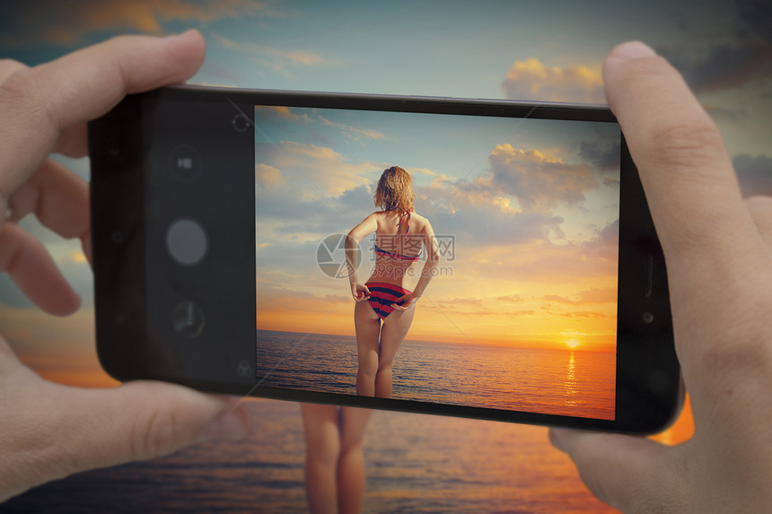 日落时,穿着泳衣的女孩站海边手机上拍摄个博主日落时,穿着泳衣的女孩站海边图片