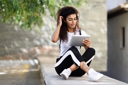 轻的北非女人看着她的数字平板电脑户外阿拉伯女孩穿着运动服城市背景图片