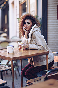 轻的阿拉伯女人坐街上的城市酒吧里阿拉伯女孩冬天穿休闲衣服,户外喝汽水背景图片