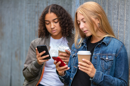 轰炸机跨种族的朋友,漂亮的金发少混血的非裔美国女孩,轻的女,用他们的智能手机社交媒体外喝外卖咖啡背景